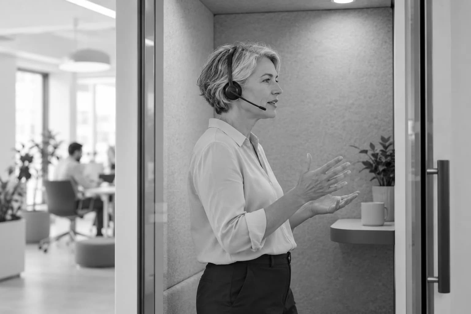 Kısa saçlı bir kadın, cam bölmeli bir ofis kabininde kulaklıkla konuşuyor.
